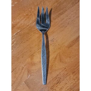May include: A stainless steel fork with three tines. The handle features an ornate, swirling pattern. The fork is designed for serving and is placed on a wooden surface.