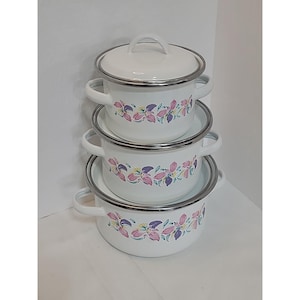 Puede incluir: Un conjunto de tres ollas de esmalte blanco con tapas y asas. Cada olla presenta un diseño floral en rosa, morado y azul. Los bordes y las asas son plateados. Las ollas están apiladas.