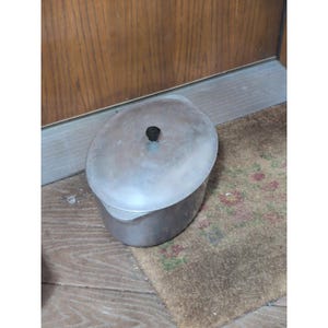 May include: A vintage, oval-shaped metal pot with a lid and a black knob. The pot is silver-colored and sits on a brown rug with floral patterns. The background includes a wooden door and a wooden floor.