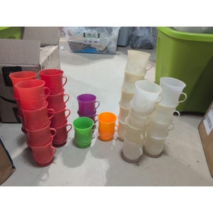May include: A selection of brightly coloured plastic mugs in shades of red, purple, green, orange, and white. The mugs are stacked in piles, some with embossed patterns. The image displays a variety of sizes and styles of these reusable drinking cups.