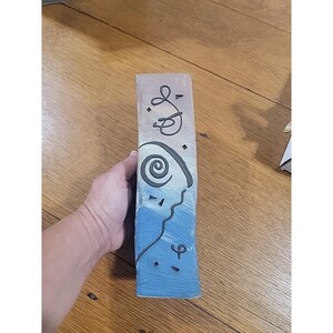 Puede incluir: Un jarrón rectangular de cerámica con la parte superior de color beige claro y la parte inferior azul. El jarrón presenta diseños abstractos negros, incluyendo una espiral, líneas curvas y pequeños puntos. El jarrón es sostenido por una mano.