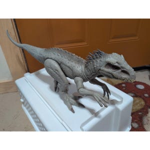 Könnte beinhalten: Eine graue Dinosaurier-Actionfigur mit strukturiertem, schuppigem Körper, scharfen Krallen und sichtbaren Zähnen. Die Figur verfügt über einen langen, spitzen Schwanz, einen stacheligen Kamm entlang des Rückens und bewegliche Gliedmaßen in Raubtierpose.