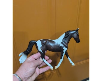 May include: A brown and white pinto horse figurine featuring a dark brown coat with white patches on the body, legs, and mane. The horse is depicted in a standing position with a long, flowing black and white tail and a sculpted muscular build.