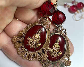 Könnte beinhalten: Ohrringe in Goldton mit roten Glasperlen und einem filigranen Design in Goldton, das einen roten Kamee mit einem goldfarbenen Bild eines sitzenden Buddha zeigt.