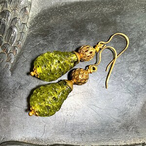 May include: A pair of gold earrings with green glass teardrop-shaped pendants. The pendants have a textured, almost honeycomb-like design.