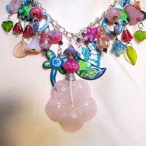 May include: A silver chain necklace with a variety of colorful charms, including flowers, leaves, and beads. The necklace features a large pink flower pendant.