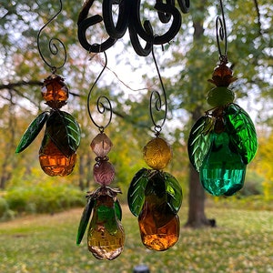 May include: Four glass ornaments with green, brown, and gold accents. Each ornament features a pear-shaped glass bead with a cluster of green leaves. The ornaments are hanging from a metal fixture.