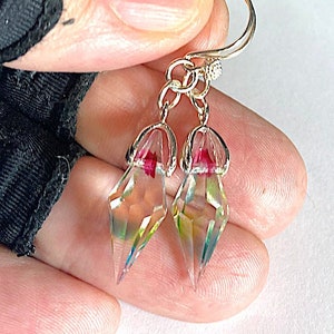 Puede incluir: Un par de pendientes de cristal iridiscente con un engaste dorado. Los cristales son facetados y tienen forma de lágrima. Los pendientes están unidos a una cadena dorada.