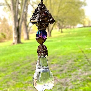 May include: A crystal teardrop suncatcher with a gold and purple beaded top. The suncatcher is hanging from a gold wire loop.
