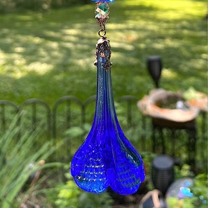 May include: A blue glass teardrop-shaped ornament with a white floral design on the top bead. The ornament is hanging from a metal chain.