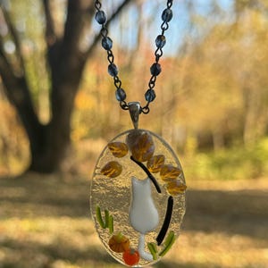 May include: A glass pendant necklace with a white cat, orange and yellow leaves, and green grass. The necklace is on a silver chain with blue beads.