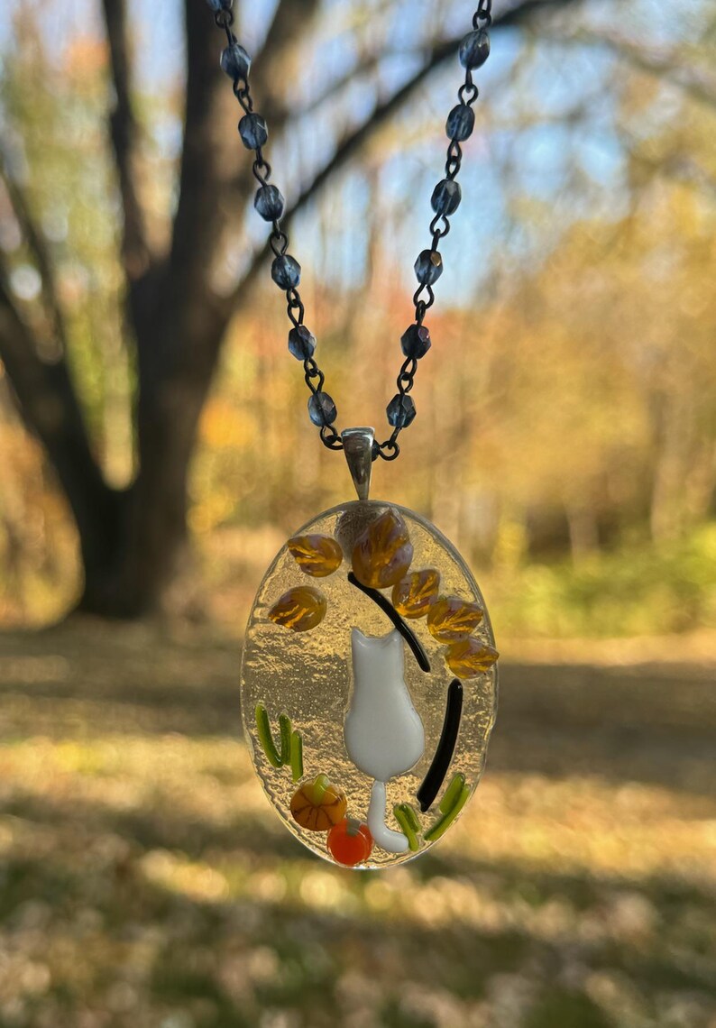 May include: A glass pendant necklace with a white cat, orange and yellow leaves, and green grass. The necklace is on a silver chain with blue beads.