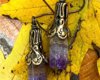 Könnte beinhalten: Ein Paar Amethyst-Kristall-Ohrringe mit verzierten goldfarbenen Metallakzenten. Die Ohrringe hängen an einem gelben Blatt.