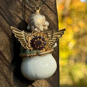 May include: A white porcelain angel ornament with gold wings and a red jeweled flower. The angel is holding a white pot.