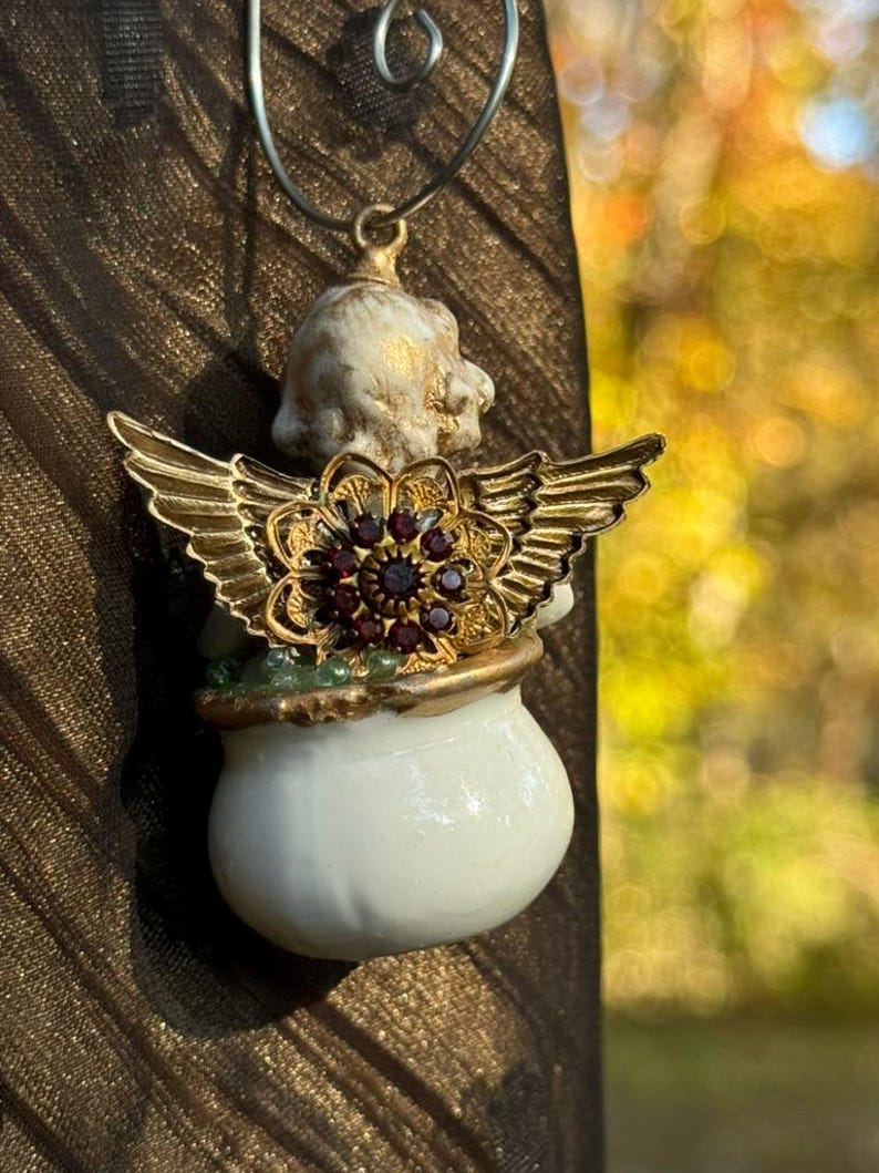 May include: A white porcelain angel ornament with gold wings and a red jeweled flower. The angel is holding a white pot.