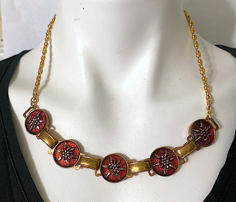May include: A gold chain necklace with five red and silver enamel pendants featuring a starburst design.