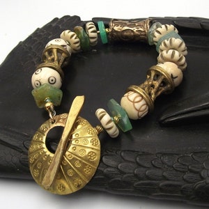 May include: A white and gold bracelet with a large, ornate gold pendant. The bracelet features a variety of beads, including bone, glass, and metal. The pendant is a circular design with a central opening and a decorative pattern.