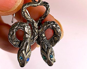 Könnte beinhalten: Silberne Schlangenohrringe mit blauen und grünen Edelsteinaugen. Die Schlangen sind ineinander verschlungen und haben ein detailliertes, strukturiertes Design.