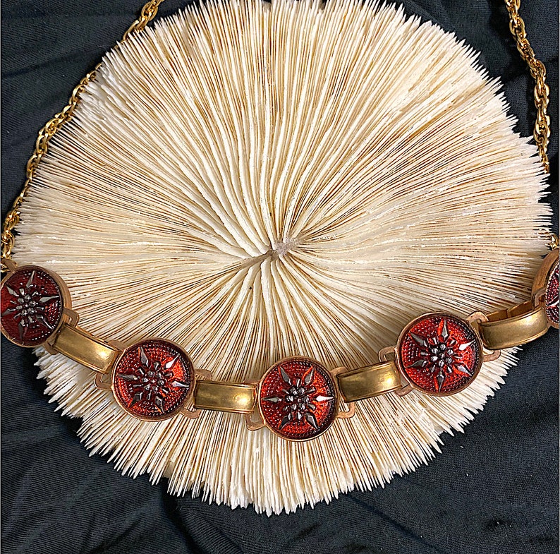 May include: A gold chain necklace with five red and gold star-shaped pendants. The necklace is lying on a white coral-like surface.