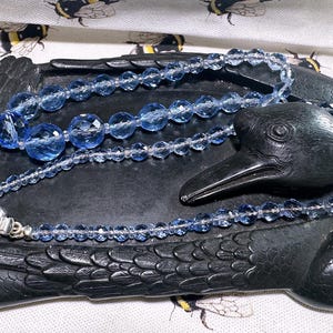 May include: A blue beaded necklace with a silver clasp rests on a black raven-shaped dish. The necklace features faceted beads of varying sizes. The dish is on a fabric with a bee pattern.