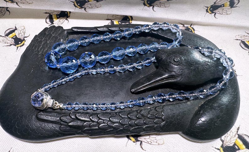 May include: A blue beaded necklace with a silver clasp rests on a black raven-shaped dish. The necklace features faceted beads of varying sizes. The dish is on a fabric with a bee pattern.