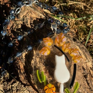 May include: A white glass cat pendant with a black tail and orange and green glass accents. The pendant is hanging from a silver chain with blue glass beads.