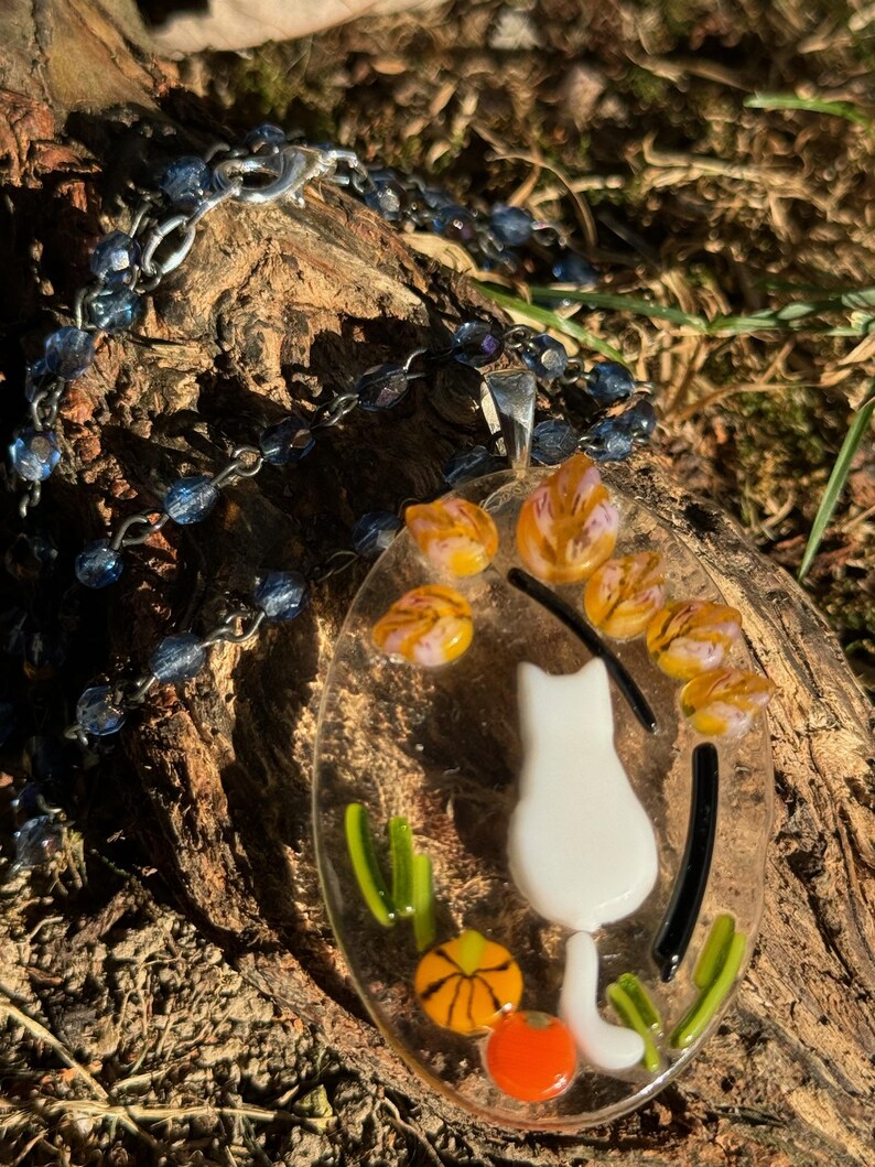 May include: A white glass cat pendant with a black tail and orange and green glass accents. The pendant is hanging from a silver chain with blue glass beads.