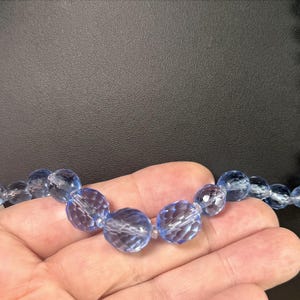 May include: A light blue faceted bead necklace. The beads are a translucent, light blue color and are strung together. The necklace is held in a hand against a black background. The beads have a sparkling, crystal-like appearance.
