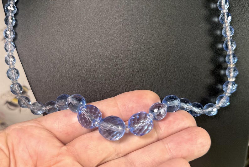 May include: A light blue faceted bead necklace. The beads are a translucent, light blue color and are strung together. The necklace is held in a hand against a black background. The beads have a sparkling, crystal-like appearance.