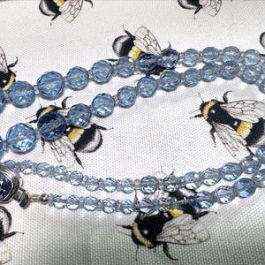 May include: A light blue beaded necklace with a silver clasp. The necklace is made of faceted beads and is displayed on a white fabric with a repeating pattern of bumblebees.