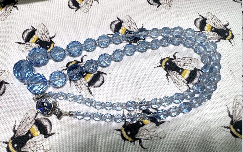 May include: A light blue beaded necklace with a silver clasp. The necklace is made of faceted beads and is displayed on a white fabric with a repeating pattern of bumblebees.