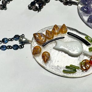 May include: A silver chain necklace with a glass pendant featuring a white cat, orange and purple flowers, and green leaves. The pendant is attached to the chain with a silver clasp.
