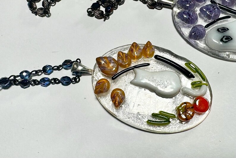 May include: A silver chain necklace with a glass pendant featuring a white cat, orange and purple flowers, and green leaves. The pendant is attached to the chain with a silver clasp.
