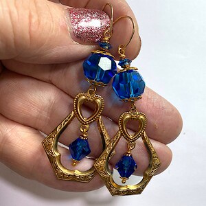 K&ouml;nnte beinhalten: Goldfarbene H&auml;ngeohrringe mit facettierten blauen Kristallperlen und herzf&ouml;rmigen Akzenten. Die Ohrringe haben ein verziertes, durchbrochenes Design und sind an goldfarbenen Ohrhaken befestigt. Die blauen Kristalle setzen einen Farbakzent auf den Schmuck im Vintage-Stil.