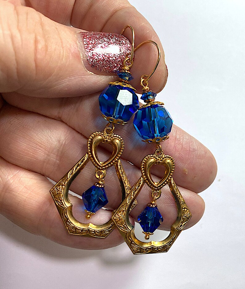K&ouml;nnte beinhalten: Goldfarbene H&auml;ngeohrringe mit facettierten blauen Kristallperlen und herzf&ouml;rmigen Akzenten. Die Ohrringe haben ein verziertes, durchbrochenes Design und sind an goldfarbenen Ohrhaken befestigt. Die blauen Kristalle setzen einen Farbakzent auf den Schmuck im Vintage-Stil.