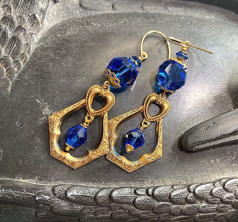 K&ouml;nnte beinhalten: Ein Paar goldfarbene Ohrringe im Vintage-Stil. Jeder Ohrring hat ein herzf&ouml;rmiges Design und einen facettierten, tiefblauen Kristall. Die Ohrringe sind etwa 7.6 cm lang und haben einen Hakenverschluss.