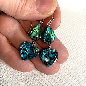 Könnte beinhalten: Ein Paar Hängeohrringe mit einem tropfenförmigen Abalone-Muschel und einer herzförmigen Abalone-Muschel. Die Ohrringe sind silberfarben und haben einen grün-blau-irisierenden Schimmer.