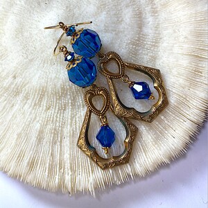 K&ouml;nnte beinhalten: Ein Paar Ohrringe im Vintage-Stil mit facettierten blauen Kristallperlen und aufwendigen goldfarbenen Metallarbeiten. Die Ohrringe haben ein Tropfendesign mit herzf&ouml;rmigen Akzenten und liegen auf einer strukturierten wei&szlig;en Oberfl&auml;che.