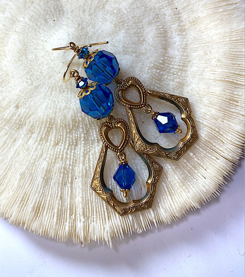 K&ouml;nnte beinhalten: Ein Paar Ohrringe im Vintage-Stil mit facettierten blauen Kristallperlen und aufwendigen goldfarbenen Metallarbeiten. Die Ohrringe haben ein Tropfendesign mit herzf&ouml;rmigen Akzenten und liegen auf einer strukturierten wei&szlig;en Oberfl&auml;che.