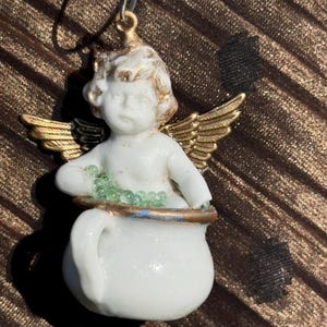 May include: A porcelain angel figurine with gold wings holding a white pot filled with green beads. The angel has a cherubic face and is wearing a white dress.