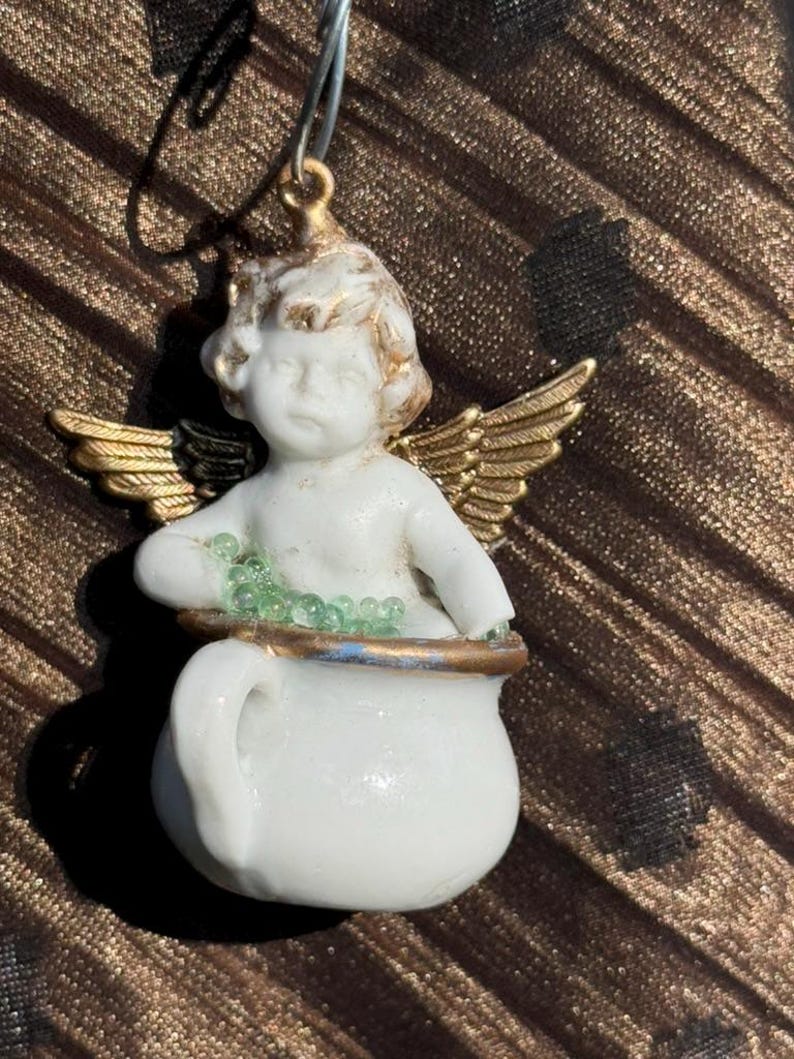 May include: A porcelain angel figurine with gold wings holding a white pot filled with green beads. The angel has a cherubic face and is wearing a white dress.