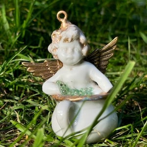 May include: A white porcelain cherub figurine with gold wings holding a pot of green beads. The cherub is sitting on a bed of green grass.