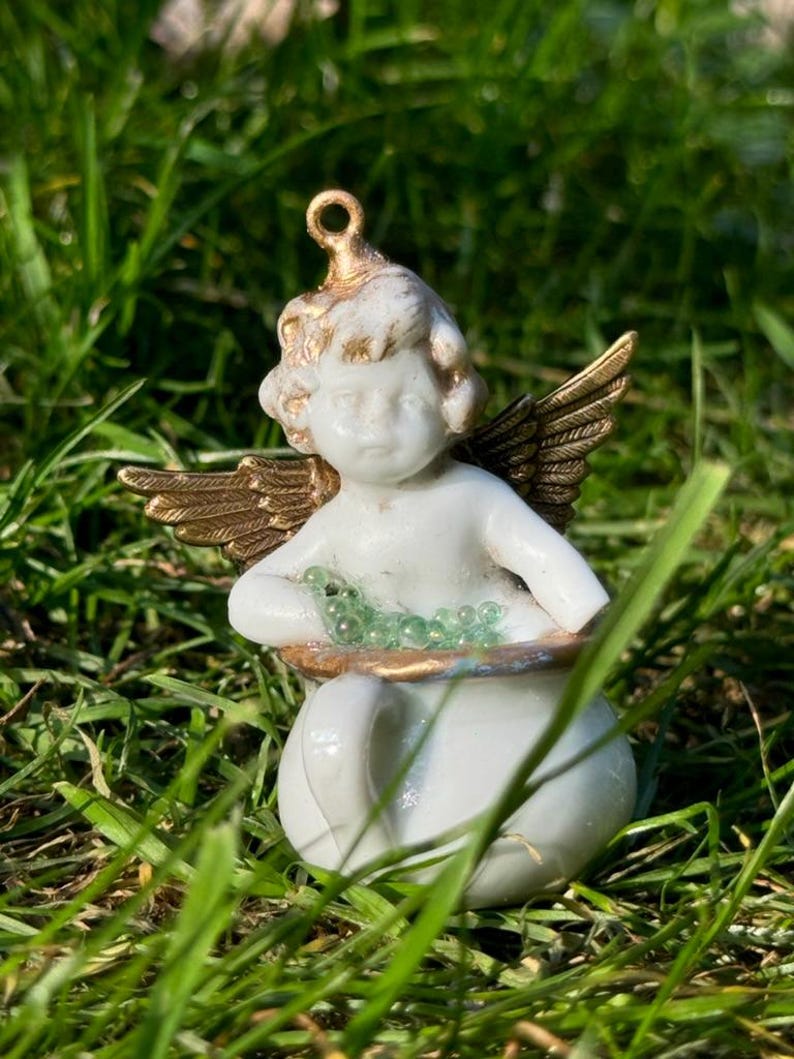 May include: A white porcelain cherub figurine with gold wings holding a pot of green beads. The cherub is sitting on a bed of green grass.