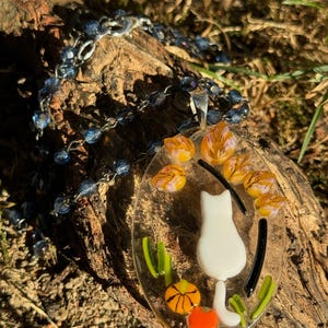 May include: A white glass cat pendant with orange and yellow flowers and a black and white striped tail. The pendant is hanging from a silver chain with blue beads.