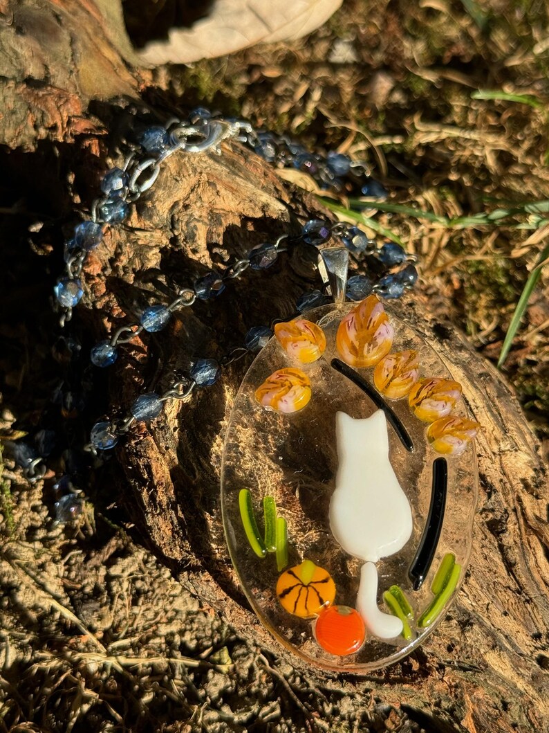 May include: A white glass cat pendant with orange and yellow flowers and a black and white striped tail. The pendant is hanging from a silver chain with blue beads.