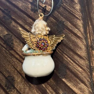 May include: A white porcelain angel ornament with gold wings and a red jeweled crown. The angel is holding a small white pot with green and gold accents.