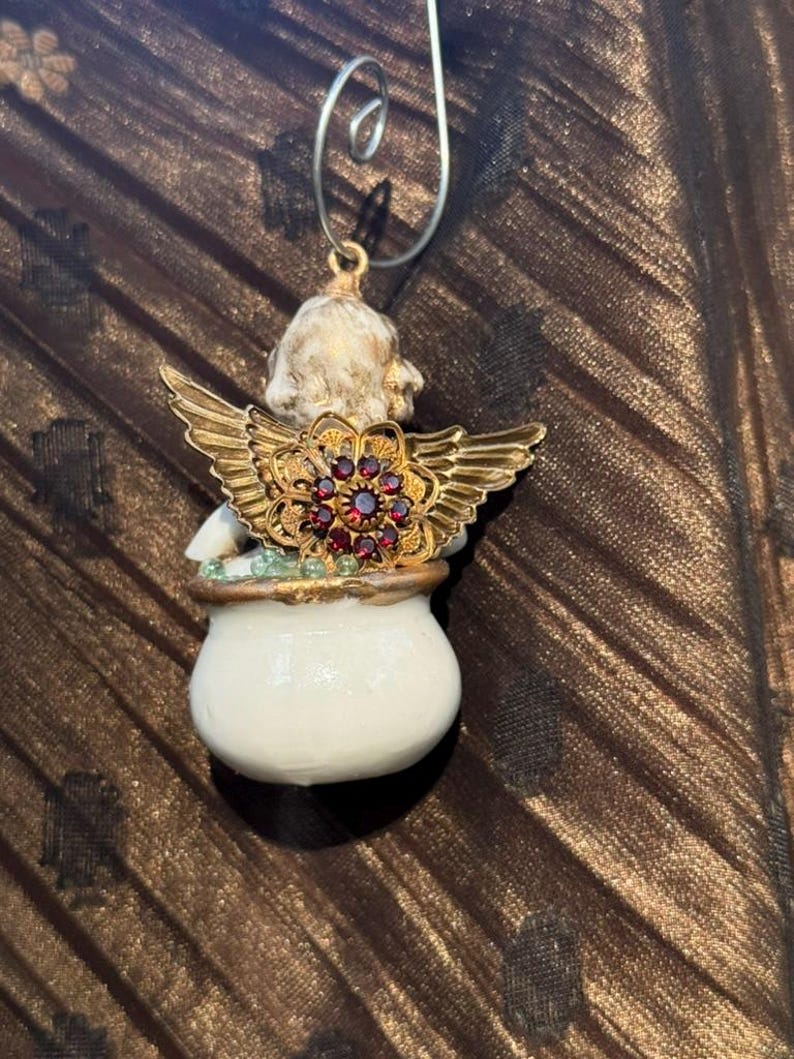May include: A white porcelain angel ornament with gold wings and a red jeweled crown. The angel is holding a small white pot with green and gold accents.