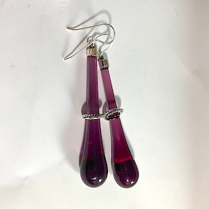 May include: A pair of dangle earrings with long, teardrop-shaped purple glass beads. The beads are attached to silver hoops with a twisted silver wire detail.