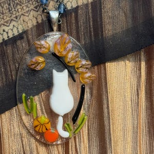 May include: A white cat with a tail curled around a pumpkin sits in a glass oval pendant necklace. The pendant is surrounded by orange, yellow, and green glass pieces.