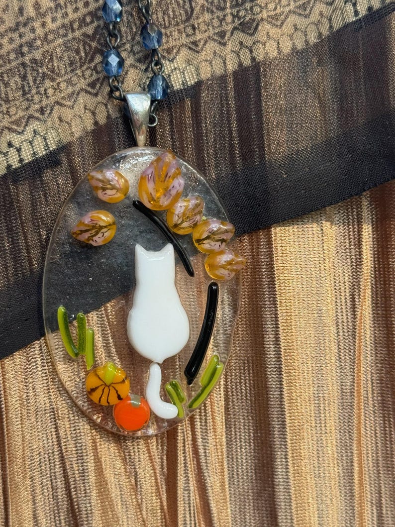 May include: A white cat with a tail curled around a pumpkin sits in a glass oval pendant necklace. The pendant is surrounded by orange, yellow, and green glass pieces.